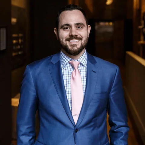 Allan Milan in a blue suit and pink tie, smiling, in a wood panelled hallway.