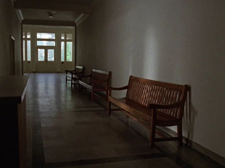 An empty corridor with three wooden benches along a pale wall and glazed doors at the far end.