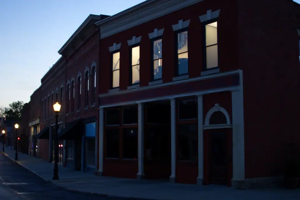 A row of brick storefronts at dusk, the shops dark and a few upstairs windows lit, iron streetlamps along an empty street.